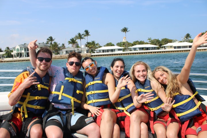 Six people in life jackets smiling and waving on a boat with palm trees in the background.