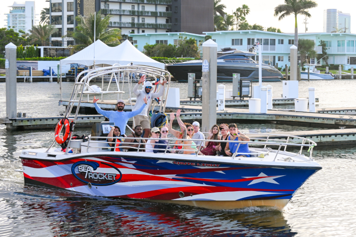 Group of people on a red, white, and blue boat in a marina waving and smiling.