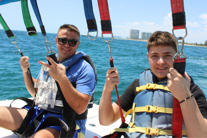 Two people parasailing over the ocean, wearing life jackets and holding onto harnesses, with a city in the background.