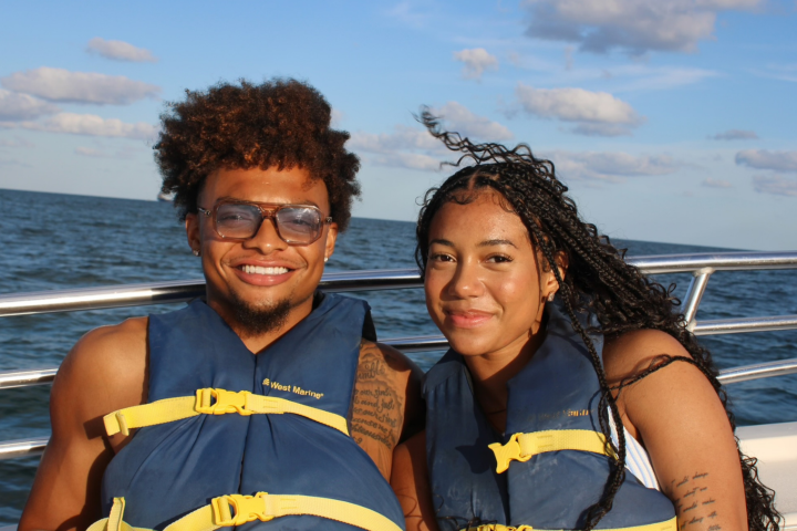 Two people wearing life vests smiling on a boat with a cloudy sky and ocean in the background.