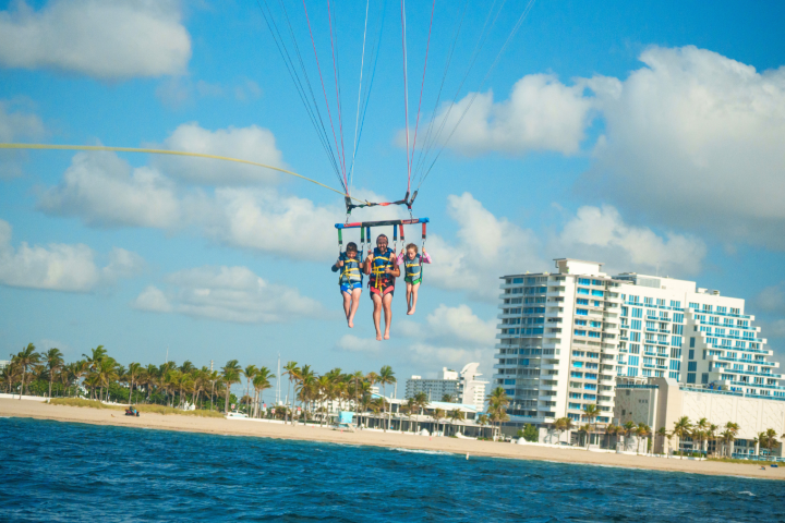 Three people parasailing above ocean near a beach with palm trees and buildings in the background.