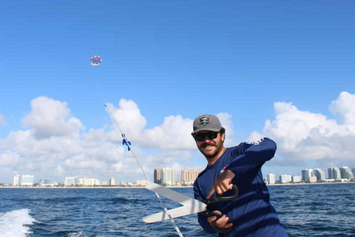 Man on boat holding a reel, parasail flying above, city skyline in background.