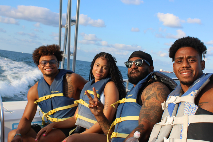 Four people on a boat wearing life jackets, smiling, with ocean in background.
