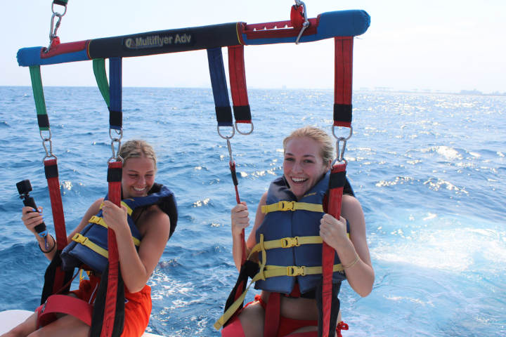 Two people parasailing over the ocean, wearing life jackets and smiling, with a clear sky in the background.