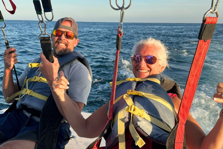 Two people parasailing over the ocean, wearing life jackets and giving thumbs up.