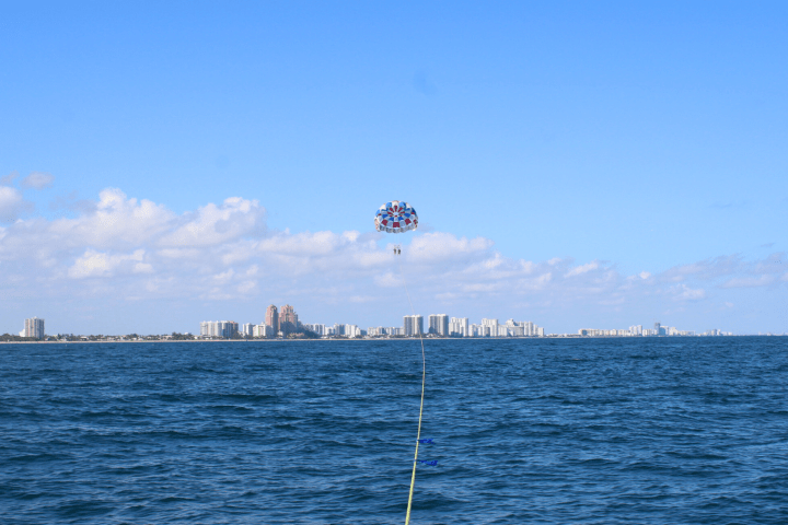 Parasail with people above ocean, city skyline in the distance under a clear blue sky.