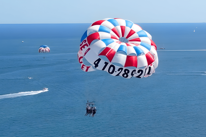 Parasailing over ocean with red, white, and blue parachute, boat and another parasailer in background.