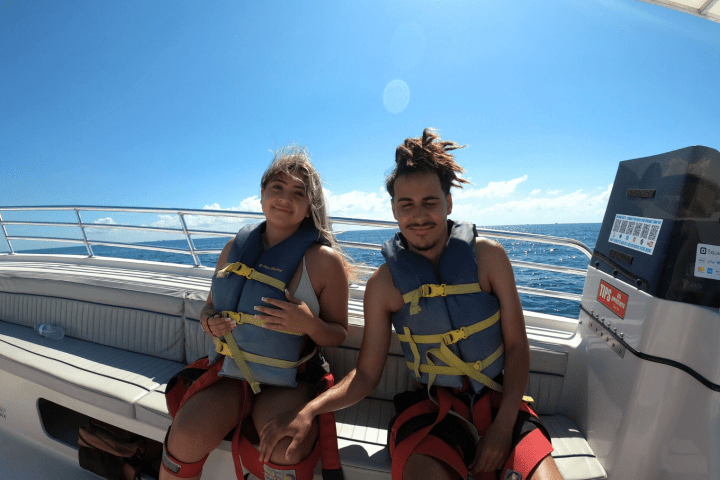 Two people wearing life jackets sitting on a boat under a bright blue sky.