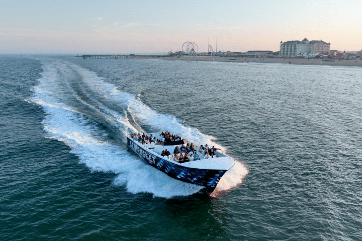 A boat with people speeds on the ocean near a coastline with buildings and a pier.