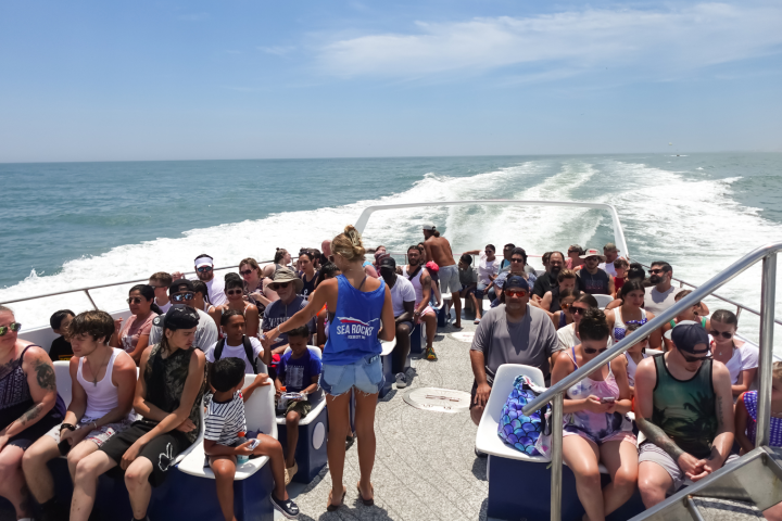 Crowded boat with people seated, ocean in the background, sunny day.