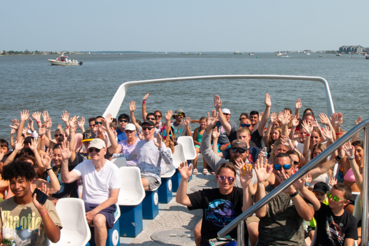 Group of people on a boat, waving, with water and other boats in the background.