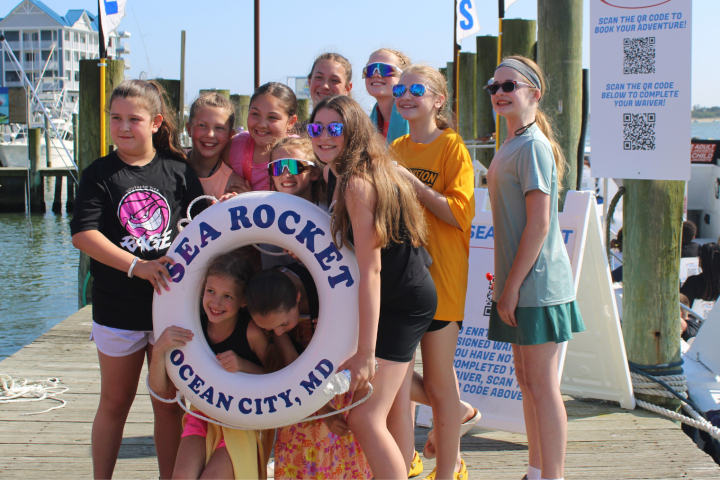 Group of children posing with a life ring on a dock, near boats.