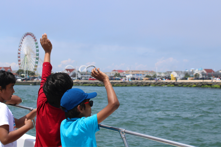 Kids on a boat wave towards a distant Ferris wheel and shoreline.
