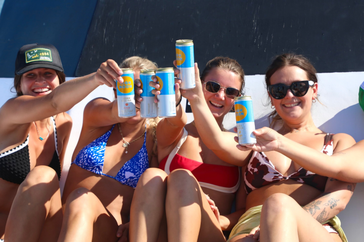 Group of women in swimwear on a boat, holding canned drinks and smiling.