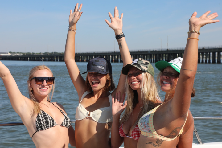 Four women in swimsuits on a boat, smiling and raising their arms.