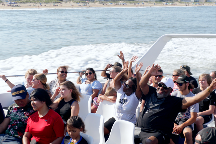 People on a boat enjoying a ride with hands raised, beach in the background.