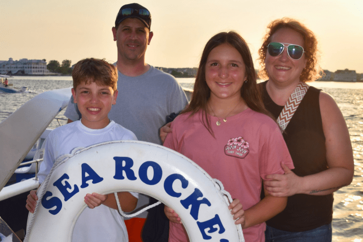 Family of four smiling on a boat holding a life ring labeled 'Sea Rocke' with sunset in the background.
