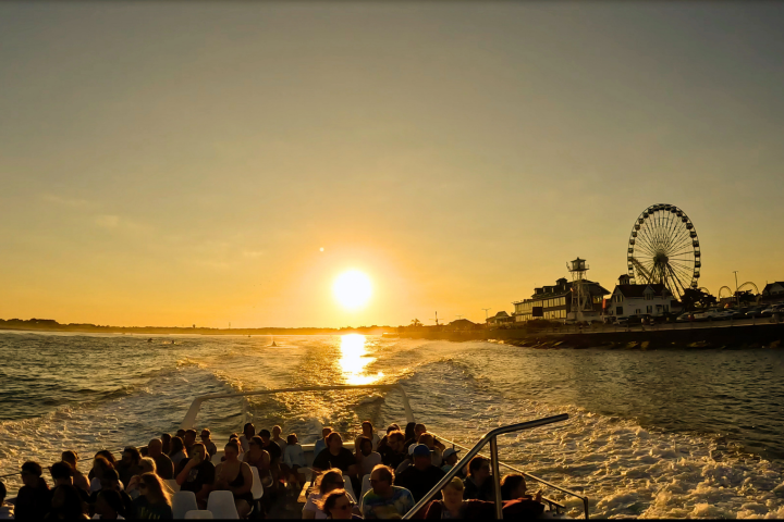 Boat with passengers at sunset near a Ferris wheel on the shoreline.