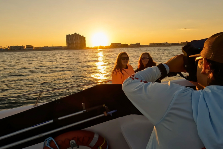 Photographer on boat takes picture of two people with sunset over water in background.