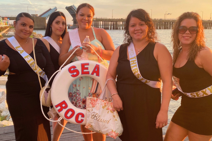 Five women in dresses with sashes stand near a lifebuoy labeled SEA ROC at sunset.