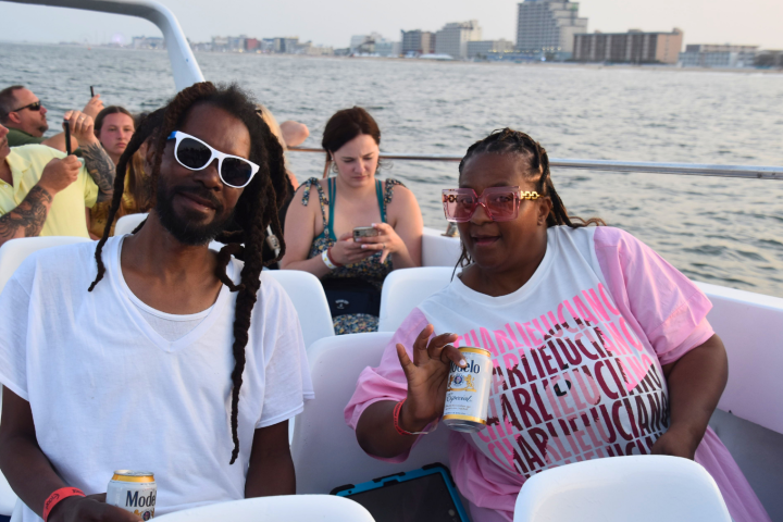 People enjoying a boat ride, two holding drinks in the foreground, city skyline in background.