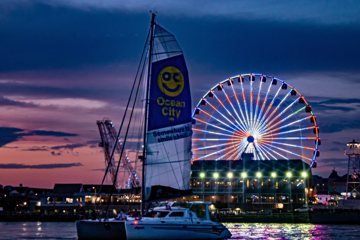 Sailboat on water at dusk with a ferris wheel lit up in the background.