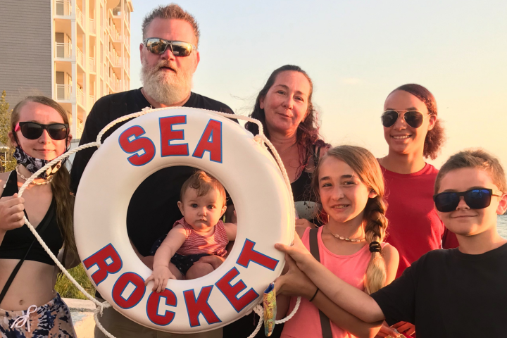 Group of people posing with a lifebuoy that reads 'SEA ROCKET' near waterfront.