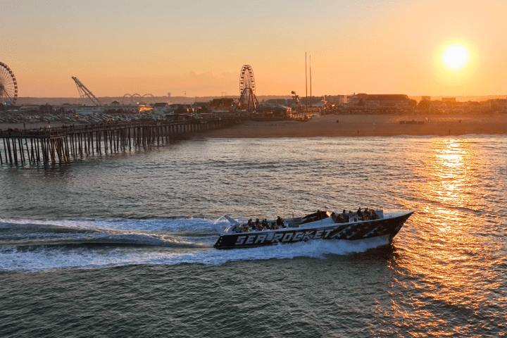 Boat speeding past pier at sunset with amusement park in background.