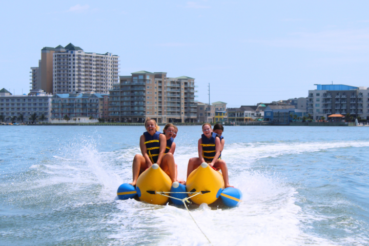 Four people riding a banana boat on the water near a city shoreline.