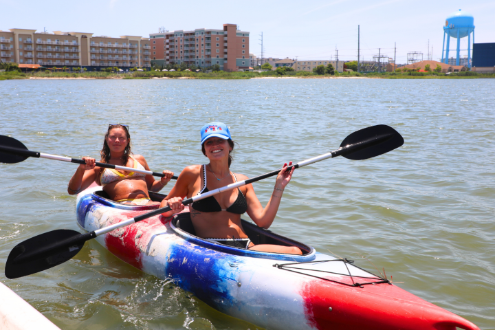 Two people kayaking on a body of water with buildings and a water tower in the background.