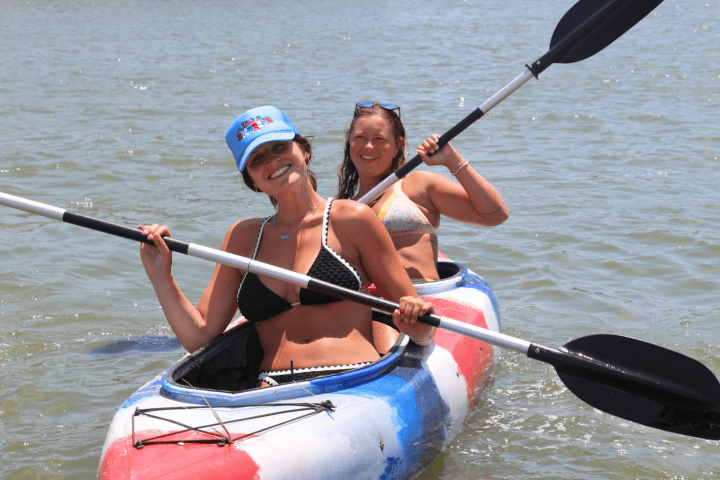 Two people kayaking on water, smiling and holding paddles, wearing swimsuits and hats.