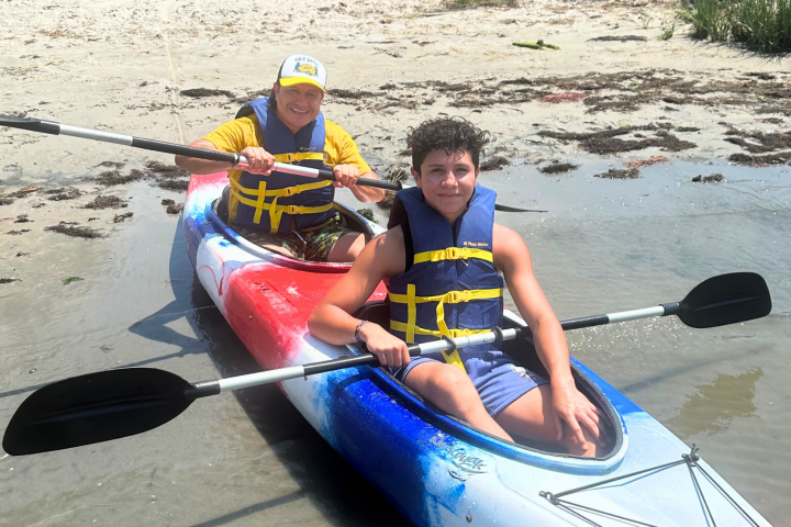 Two people in life vests kayaking on a calm beach.