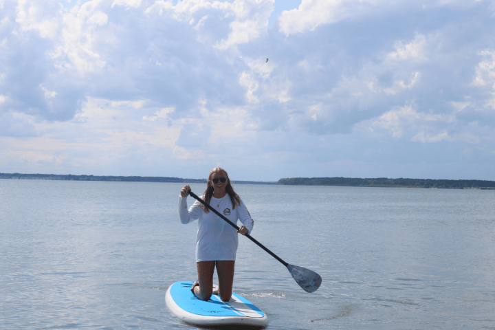 Person kneeling on a paddleboard with a paddle on a calm lake.