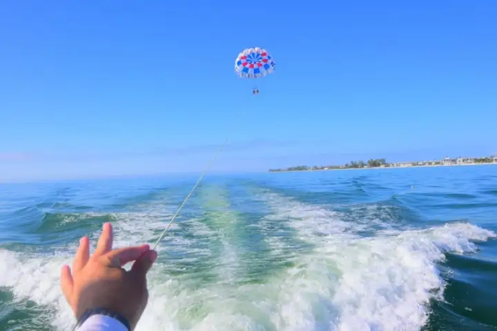 Hand pointing at a parasailer over the ocean with a clear blue sky and distant shoreline.