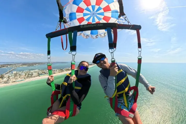 Two people parasailing above green water and beach with a colorful parachute under clear blue sky.