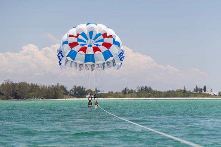 Two people parasailing over turquoise water with a red, white, and blue parachute near a shoreline.