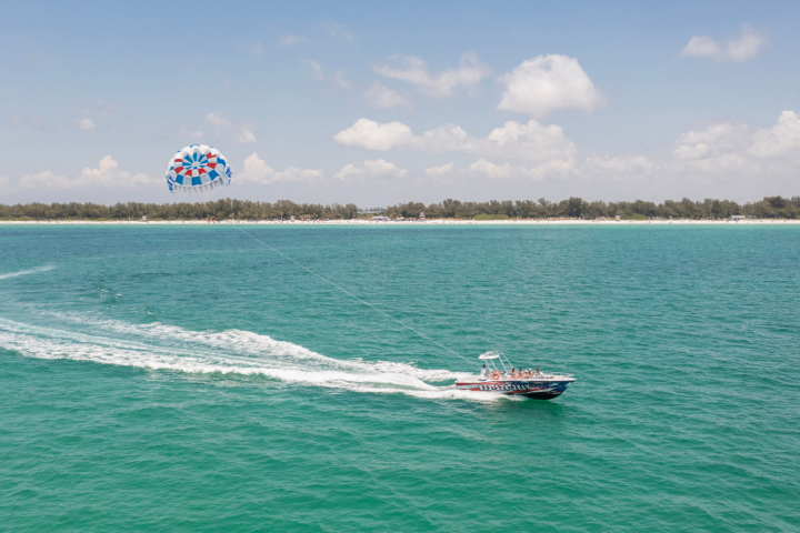 Boat towing a parasail over turquoise water with a distant shoreline and cloudy sky.