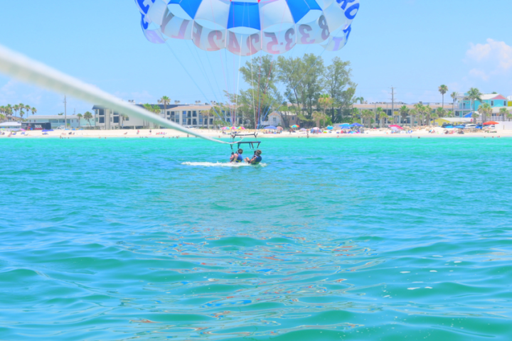 Parasailing over turquoise water near a sandy beach with buildings and palm trees.