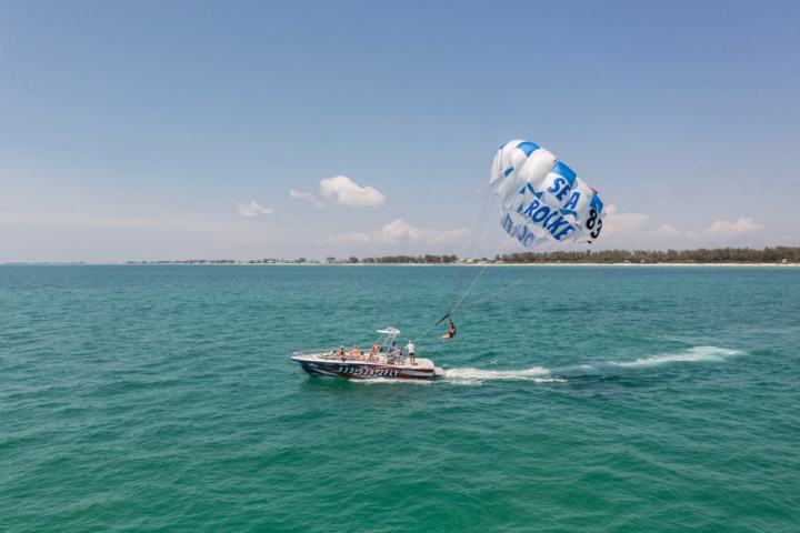Boat towing a parasail with a person over blue ocean water near the shoreline on a clear day.