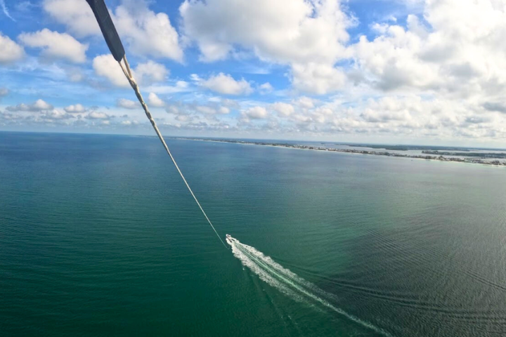Aerial view of a boat pulling a parasail over open water with a coastline in the distance.