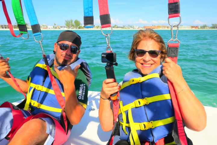Two people with sunglasses and life vests sit on a boat, smiling over turquoise water.