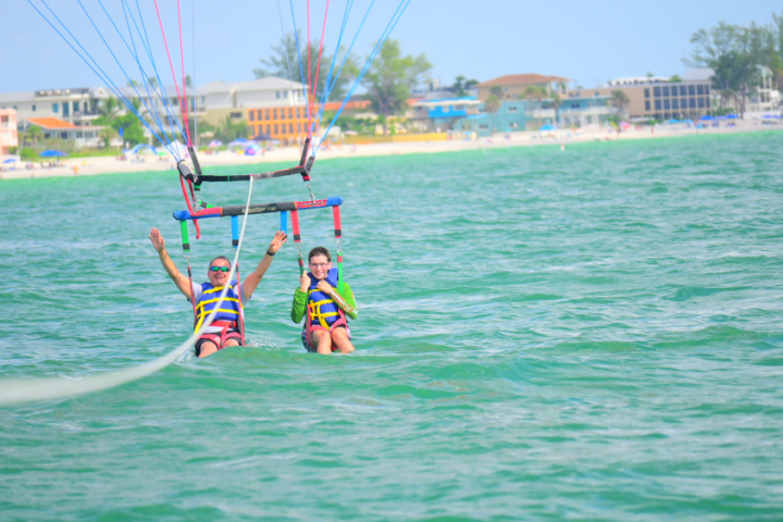 Two people parasailing over the ocean near a beach with buildings in the background.