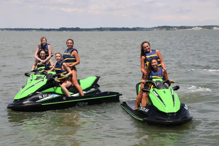 Three green jet skis with six people on a lake, wearing life jackets.
