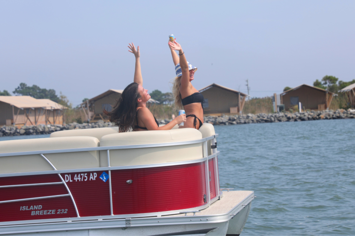 Two people on a red pontoon boat, enjoying the day and raising arms, with scenic shoreline in the background.