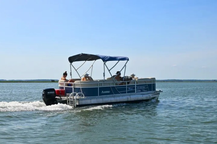 Pontoon boat with several people cruising on a calm lake under a clear blue sky.