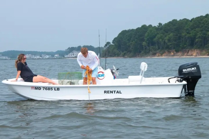 Two people on a small rental boat over calm water, one sitting and one handling fishing gear.