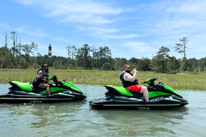 Two people riding green jet skis on a river with trees and a distant lighthouse.