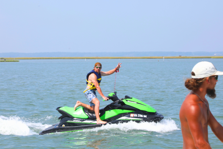Man on green jet ski in water, another man walking on shore.
