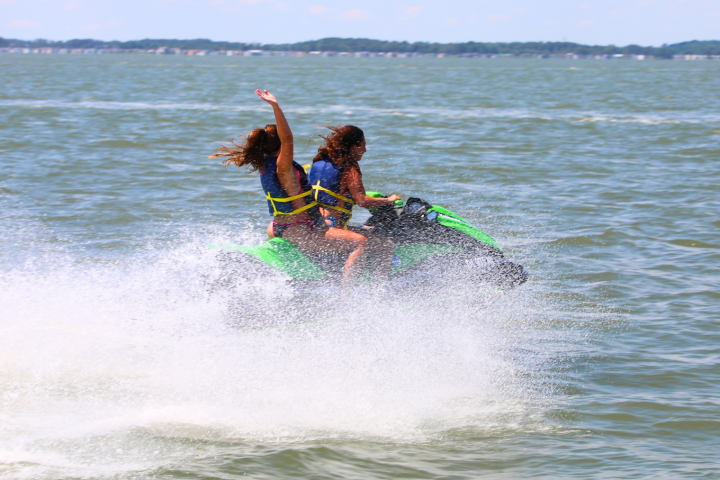 Two people on a jet ski, wearing life jackets, speeding on a lake.