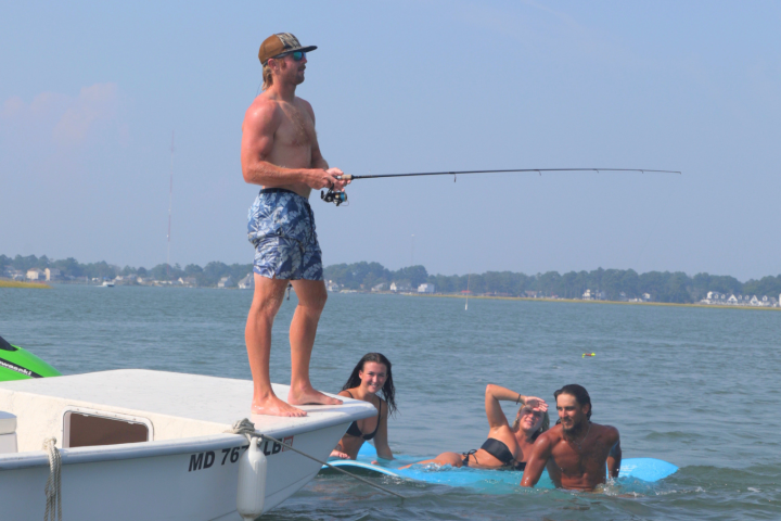 Man fishing from boat while three people float nearby in water.